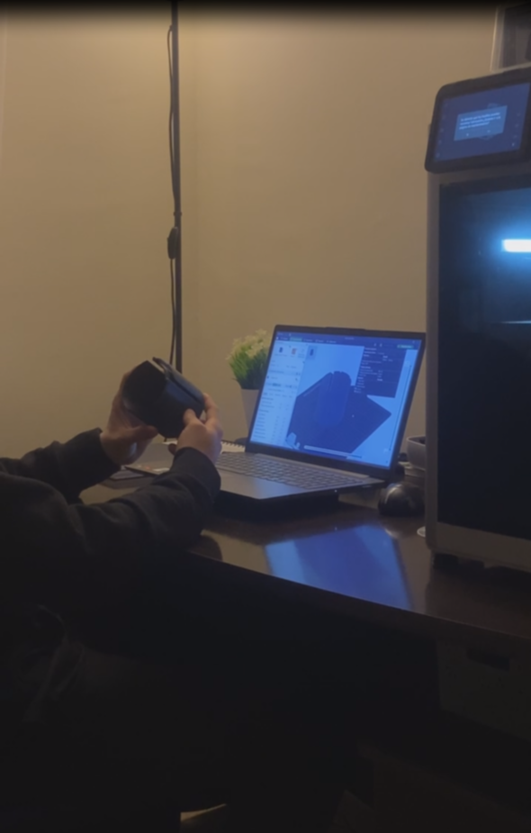 Persona sosteniendo una pieza impresa en 3D junto al portátil con el diseño y la impresora 3D del taller
